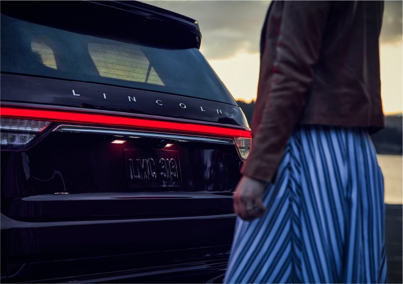 A person is shown near the rear of a 2023 Lincoln Aviator® SUV as the Lincoln Embrace illuminates the rear lights | Palmetto Lincoln in Charleston SC