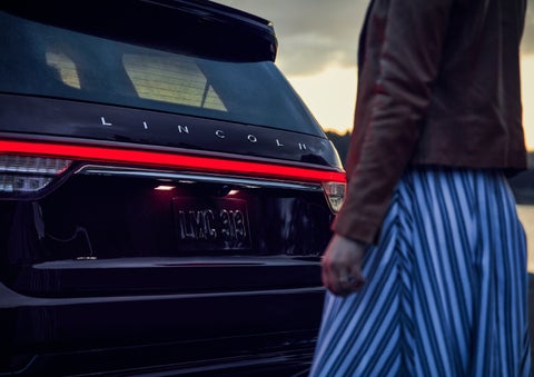 A person is shown near the rear of a 2024 Lincoln Aviator® SUV as the Lincoln Embrace illuminates the rear lights | Palmetto Lincoln in Charleston SC